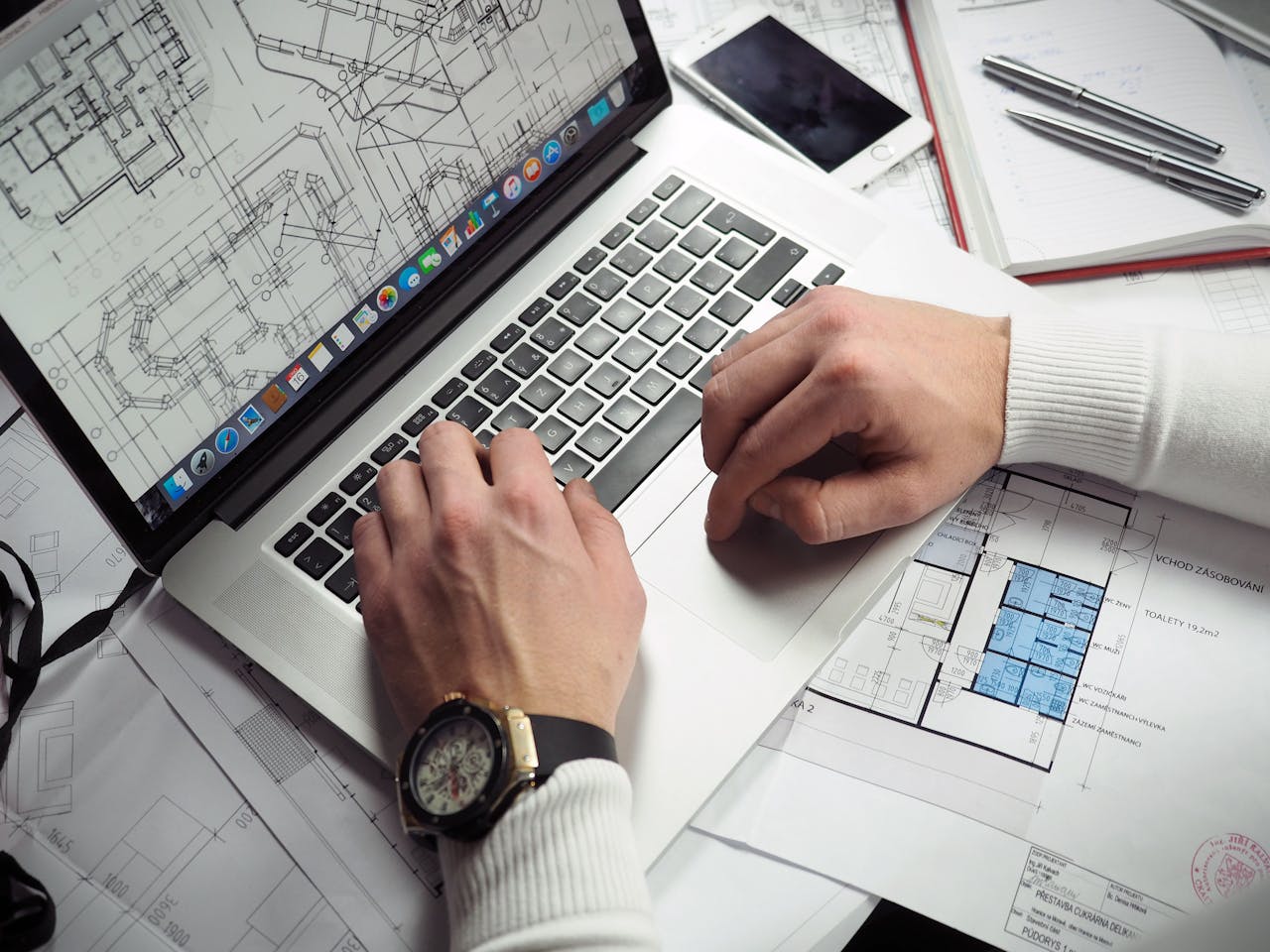 services-04 Architect reviewing detailed floor plans and schematics on a laptop with a smartphone nearby.