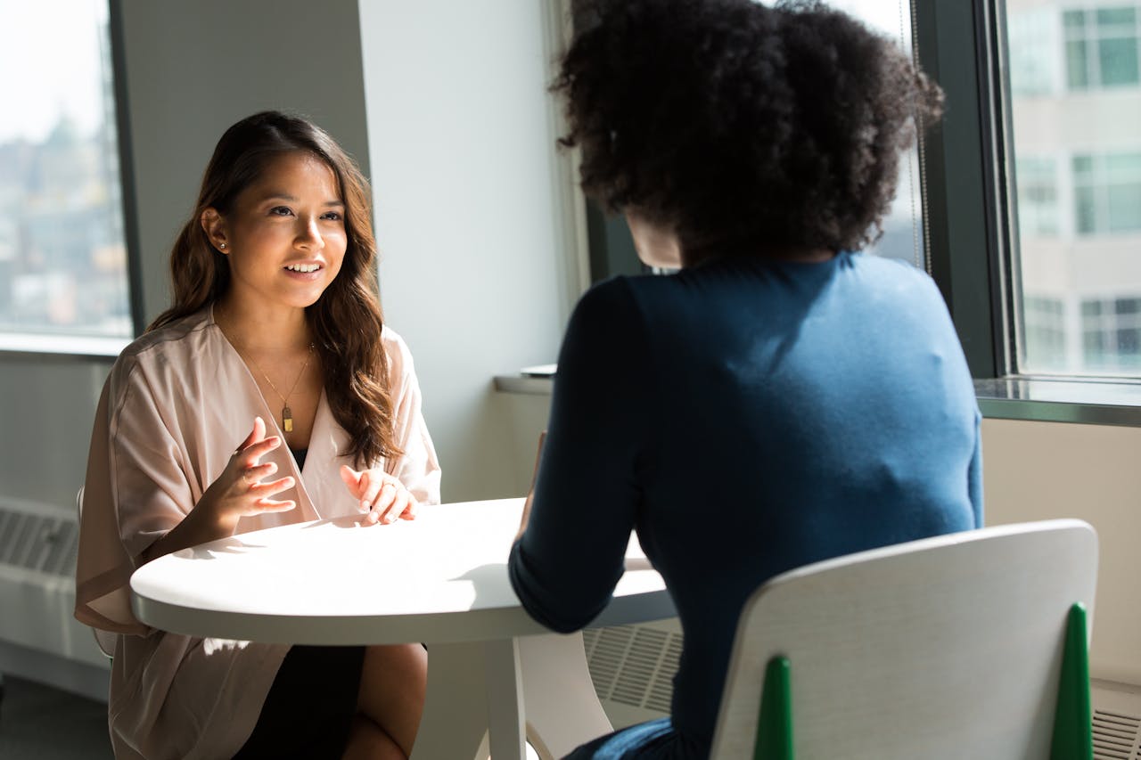 why-choose-us Two women sitting at a table having a professional discussion in a bright office setting.