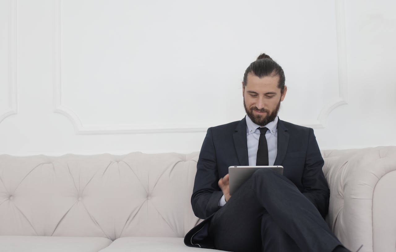 about-us A businessman in a suit sitting on a sofa using a tablet, smiling.